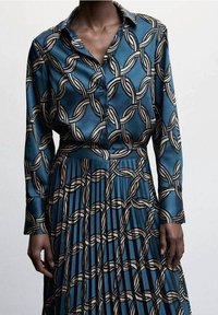 Ensemble chemisier en soie et jupe couleur sarcelle, avec un motif chaîne en crème et noir. Des plis texturés sur la jupe ajoutent un intérêt visuel.