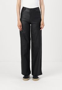 Pantalons larges noirs en simili cuir, avec une taille haute, des poches avant et des détails de couture visibles. Associés à des baskets noires.