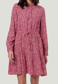 Femme portant une robe longue à manches longues rouge avec un imprimé floral blanc, devant boutonnée et avec une taille à cordon, se tenant contre un fond gris clair.