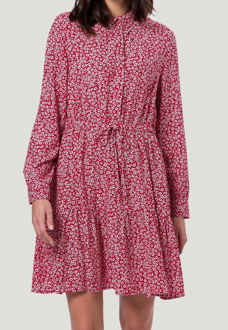 Femme portant une robe longue à manches longues rouge avec un imprimé floral blanc, devant boutonnée et avec une taille à cordon, se tenant contre un fond gris clair.
