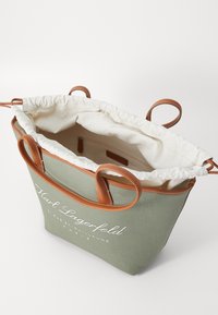 Grønn canvas tote bag med brune lærdetaljer og håndtak, med snøring og hvit logo i front.