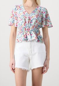 Blusa floreale con scollo a V, maniche corte e vita arricciata in azzurro chiaro, con motivi rosa, gialli e verdi. Indossata con pantaloni corti bianchi sfilacciati.