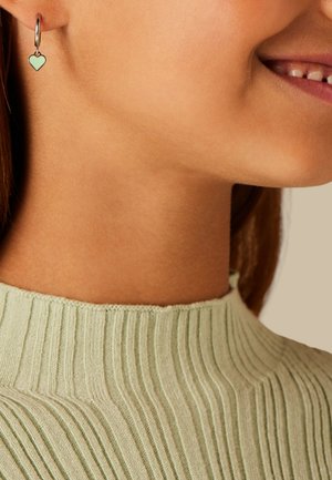 Nahaufnahme einer lächelnden Person, die einen hellgrünen, gerippten Pullover und einen silbernen Creolen-Ohrring mit einem mintgrünen Herzanhänger trägt.