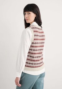 Adīta veste ar rakstainu dizainu bordo, krēmkrāsas un tumši sarkanā tonī. Iespējo ribota apmale un apaļa kakla izgriezuma forma, valkāta virs baltas krekla.