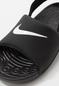 Musta liukumaton sandaali matalla pinnalla, jossa on silmiinpistävä valkoinen Nike-logo, teksturoitu musta pohja pyöreillä kuvioilla ja säädettävä hihna.