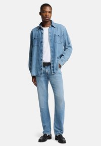 Helles blaues Denimhemd mit zwei Brusttaschen, kombiniert mit einem weißen T-Shirt und hellen Denim-Jeans. Der schwarze Gürtel und die Schuhe runden das Outfit ab.