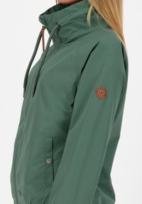 Grøn vandtæt jakke med høj krave, snøre, sidelommer og et rundt brunt logo-patch på ærmet. Glat tekstur.