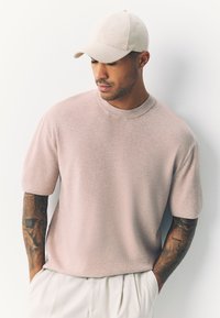 Ljusrosa kortärmad stickad tröja med rund halsringning, bärs med en beige keps. Mannens bara arm har detaljerade tatueringar.