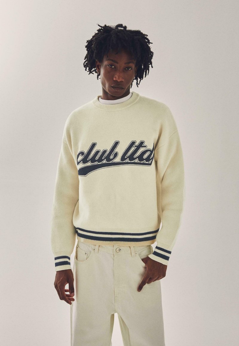 Uomo con capelli ricci che indossa un maglione crema con la scritta "club ltd." e pantaloni crema coordinati, in piedi davanti a uno sfondo chiaro e semplice.