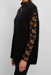 Gilet nero a maglia con scollatura a V profonda e ampie aperture per le braccia, indossato sopra una camicia floreale scura con accenti arancioni. Tessuto texture, vestibilità rilassata.
