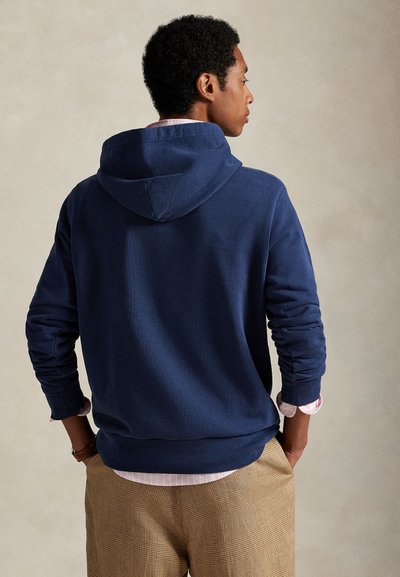 Bărbat purtând hanorac bleumarin, cămașă roz deschis și pantaloni în carouri bej, stând cu mâinile în buzunare, față în profil pe fundal simplu.