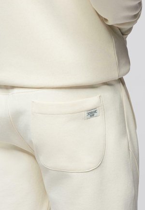 Detailní záběr osoby nosící béžové tepláky se zadní kapsou a malou obdélníkovou látkovou cedulkou připevněnou na ní.