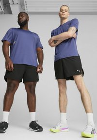 Due modelli maschili stanno fianco a fianco, indossando camicie viola e pantaloni corti neri. Uno indossa sneakers nere, l'altro ha scarpe bianche e neon.