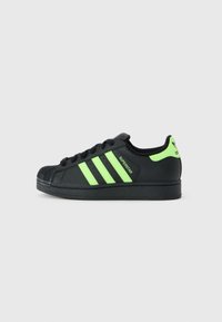 Baskets Adidas Superstar noires avec des rayures vert fluo, un onglet au talon et le texte "Superstar" sur le côté, présentées de l'extérieur.