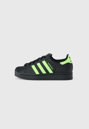 Baskets Adidas Superstar noires avec des rayures vert fluo, un onglet au talon et le texte "Superstar" sur le côté, présentées de l'extérieur.