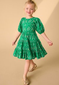 Abito floreale verde con maniche a palloncino, gonna a strati e sandali dorati. Il tessuto presenta un motivo di fiori e foglie.
