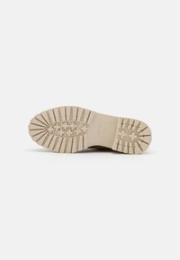 Suela de zapatilla en goma beige claro con un patrón de superficie texturizado. Presenta orificios circulares para la transpirabilidad y una construcción sólida.