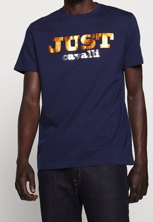 Mężczyzna noszący granatowy t-shirt z metalicznym złotym napisem "JUST" i srebrnym "cavalli" na piersi, w połączeniu z ciemnymi jeansami.