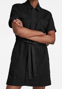 Person med korslagda armar som bär en kortärmad svart klänning med bälte, knappade bröstfickor och krage.