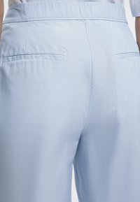 Οπίσθια όψη ανοιχτόμπλε καλοραμμένων παντελονιών με δύο οριζόντιες τσέπες και εφαρμοστό ζωνάρι σε άτομο που φοράει λευκό πουκάμισο.