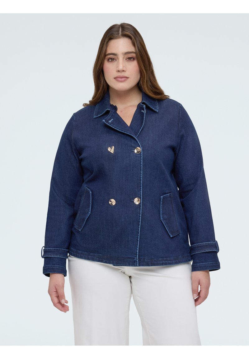 Donna che indossa una giacca di jeans blu scuro con bottoni dorati e pantaloni bianchi, in piedi davanti a uno sfondo chiaro e uniforme.