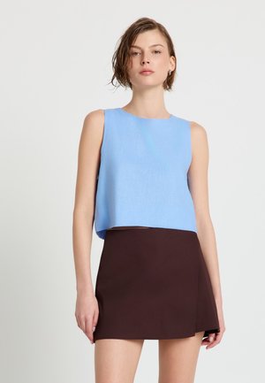 Donna con capelli corti e bagnati che indossa una canottiera azzurro chiaro e una minigonna marrone scuro, in piedi davanti a uno sfondo bianco semplice.