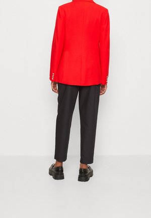 Personne portant un blazer rouge vif avec des boutons argentés sur les manches, un pantalon noir court, et des mocassins noirs épais, debout devant un fond blanc.