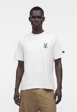 Ung mand iført en hvid t-shirt med en lille Bugs Bunny-grafik på brystet og beige bukser, der står foran en ensfarvet baggrund.