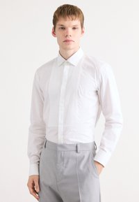 Chemise blanche à devant plissé, col classique et manches longues. Le tissu semble lisse et frais, associé à un pantalon gris clair.