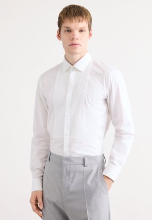 Camisa blanca de vestir con frente plegado, cuello clásico y mangas largas. La tela parece suave y fresca, combinada con pantalones gris claro.