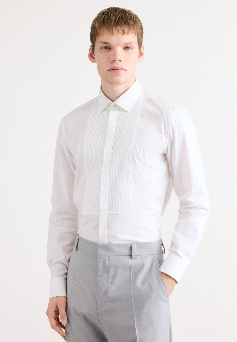 Chemise blanche à devant plissé, col classique et manches longues. Le tissu semble lisse et frais, associé à un pantalon gris clair.