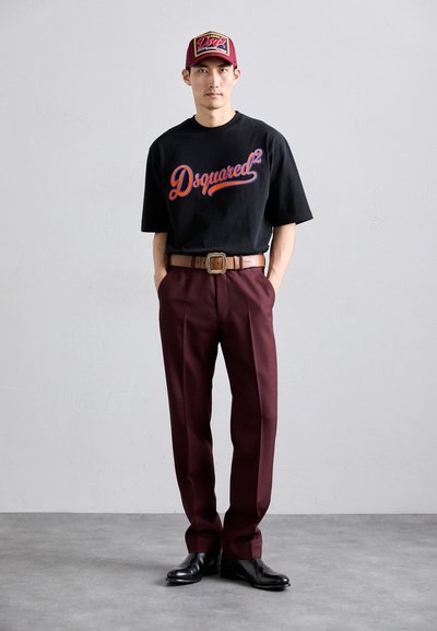 Man wearing a black Dsquared2 t-shirt, burgundy pants with a brown belt, black shoes, and a burgundy cap in a plain room.