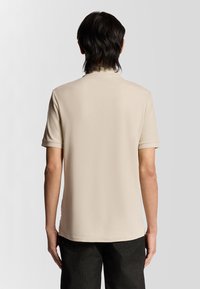 Beige polotröja med korta ärmar, slät textur och rundad krage. Baksidan har en diskret logotyp nära nacken.