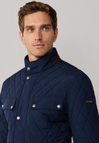 Marineblå quiltet jakke med høj krave, udstyret med sølvfarvede trykknapper og to brystlommer. Brune detaljer nær kraven.