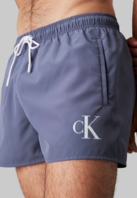 Shorts da nuoto corti di colore grigio scuro uniforme, con un cordino bianco, tasche laterali e un logo "ck" bianco sulla gamba sinistra.