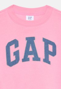 Rosa genser med rund hals. Har et stort blått denim "GAP"-logo med kontraststing. Myk tekstur, uformelt design.