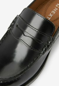 Mocassin en cuir noir avec une finition brillante, présentant des coutures décoratives, un détail de porte-monnaie et un design à bout rond lisse.