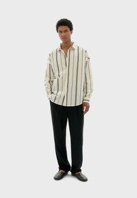 Camisa de manga larga a rayas en tela crema y negra con cierre de botones, combinada con pantalones negros y zapatillas oscuras sobre un fondo neutro.