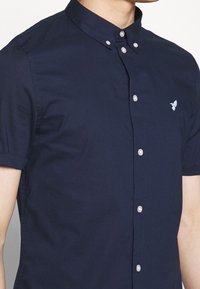 Uomo che indossa una camicia a maniche corte blu navy con un logo di un uccello sul lato sinistro del petto e colletto a bottoni.