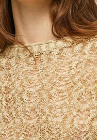 Spitzenstrickoberteil in Beige und Gold mit rundem Ausschnitt und aufwendigen offenen Mustern, das ein zartes, strukturiertes Aussehen betont.
