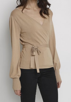 Nainen pukeutuneena beigeen kietaisuneuleeseen, jossa on solmittu vyötärö ja pussimaiset hihat, yhdistettynä mustiin housuihin yksinkertaisen taustan edessä.