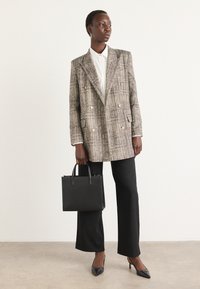 Doppelt geschnittener Blazer in strukturiertem hellbraun-grauem Muster, kombiniert mit schwarzen Hosen und schmalen Absatzschuhen, während sie eine kleine schwarze Tasche hält.