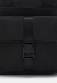 Zaino imbottito nero con chiusura a fibbia in plastica e spallacci in tessuto. La superficie è liscia con una finitura opaca e un design minimale.