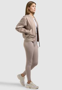 Kvinde stående i profil iført en beige satin bomberjakke, lysebeige leggings, hvid crop top og hvide træningssneakers på grå baggrund.