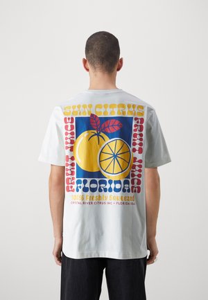 Mężczyzna w białej koszulce z kolorową grafiką całej i pokrojonej pomarańczy, liści oraz tekstem o soku z cytrusów Florydy na plecach.
