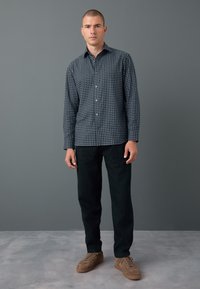 Camicia a quadri a bottoni in navy e bianco, abbinata a pantaloni scuri e scarpe in camoscio marrone. Ambientata su uno sfondo grigio.