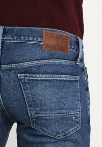 Modré džínové kalhoty s koženou značkou. Mají dvě zadní kapsy a zesílené švy. Látka je s hladkým, texturovaným povrchem.