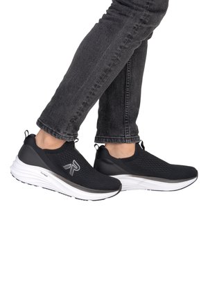 Personne portant des baskets slip-on en maille noire avec des semelles blanches et un jean noir, debout les jambes croisées sur un fond blanc.