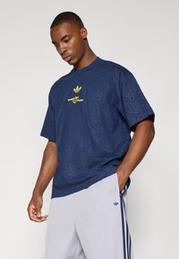 Camiseta azul marino con un diseño texturizado y estampado, que presenta un logo amarillo en el centro. Manga corta, corte relajado.