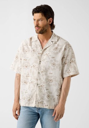 Homme aux cheveux foncés et à la barbe portant une chemise beige à manches courtes avec des motifs floraux marron et un jean bleu clair, debout et regardant vers la gauche.
